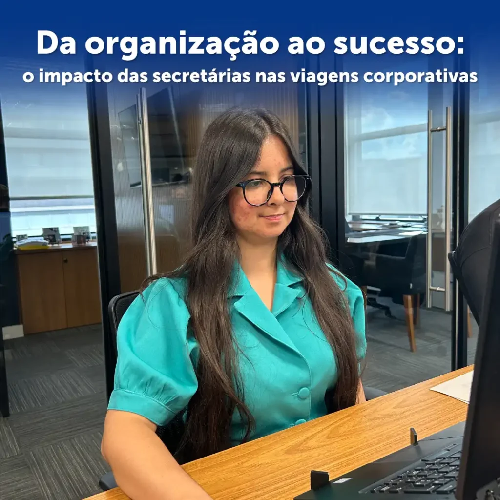 Mulher com camiseta verde água, está sentada. E em sua frente há uma mesa com computador simulando a rotina de uma secretária.