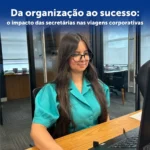 Mulher com camiseta verde água, está sentada. E em sua frente há uma mesa com computador simulando a rotina de uma secretária.